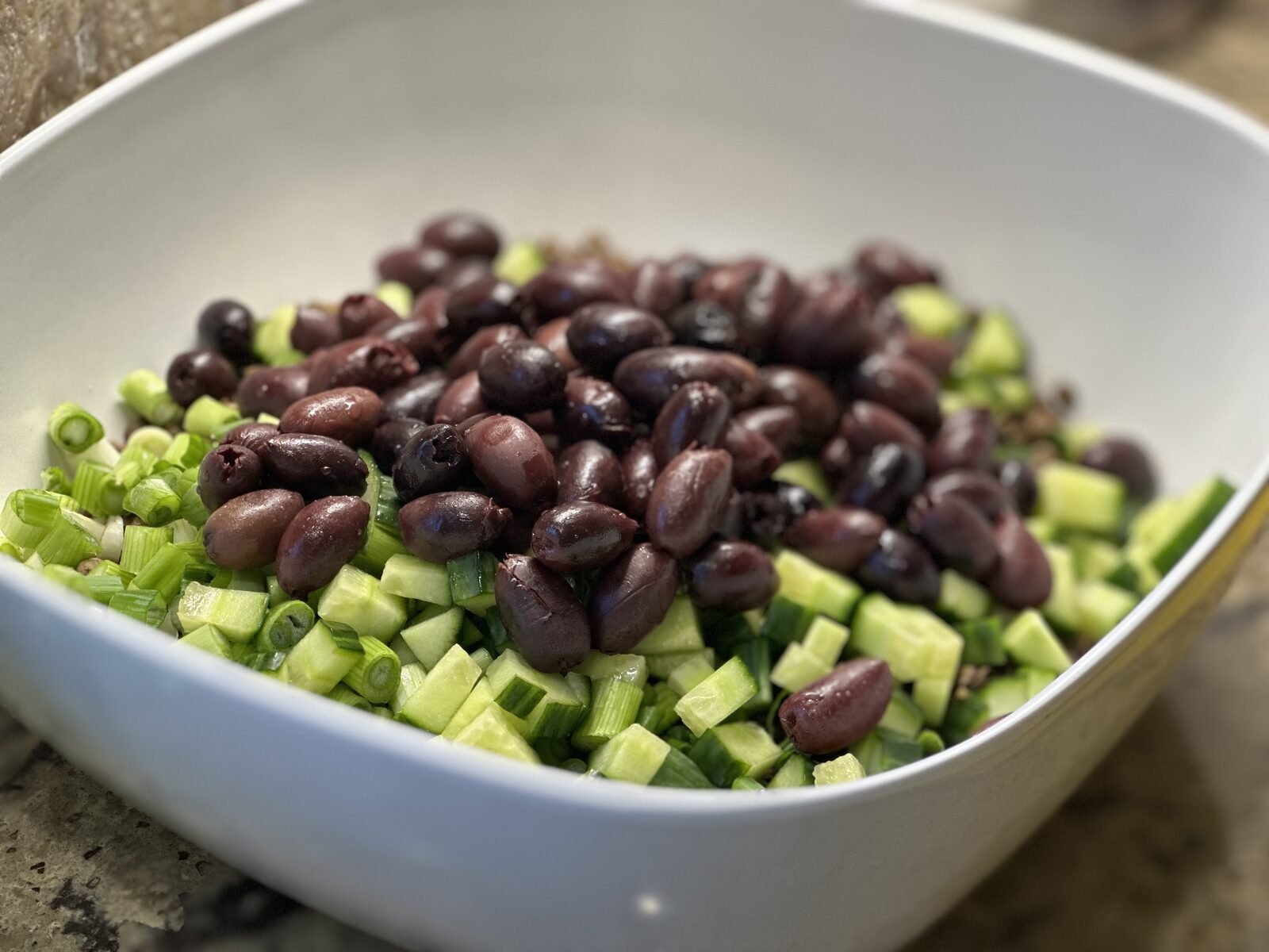 A white bowl filled with quinoa, kalamata olives, diced cucumber, and chopped scallions
