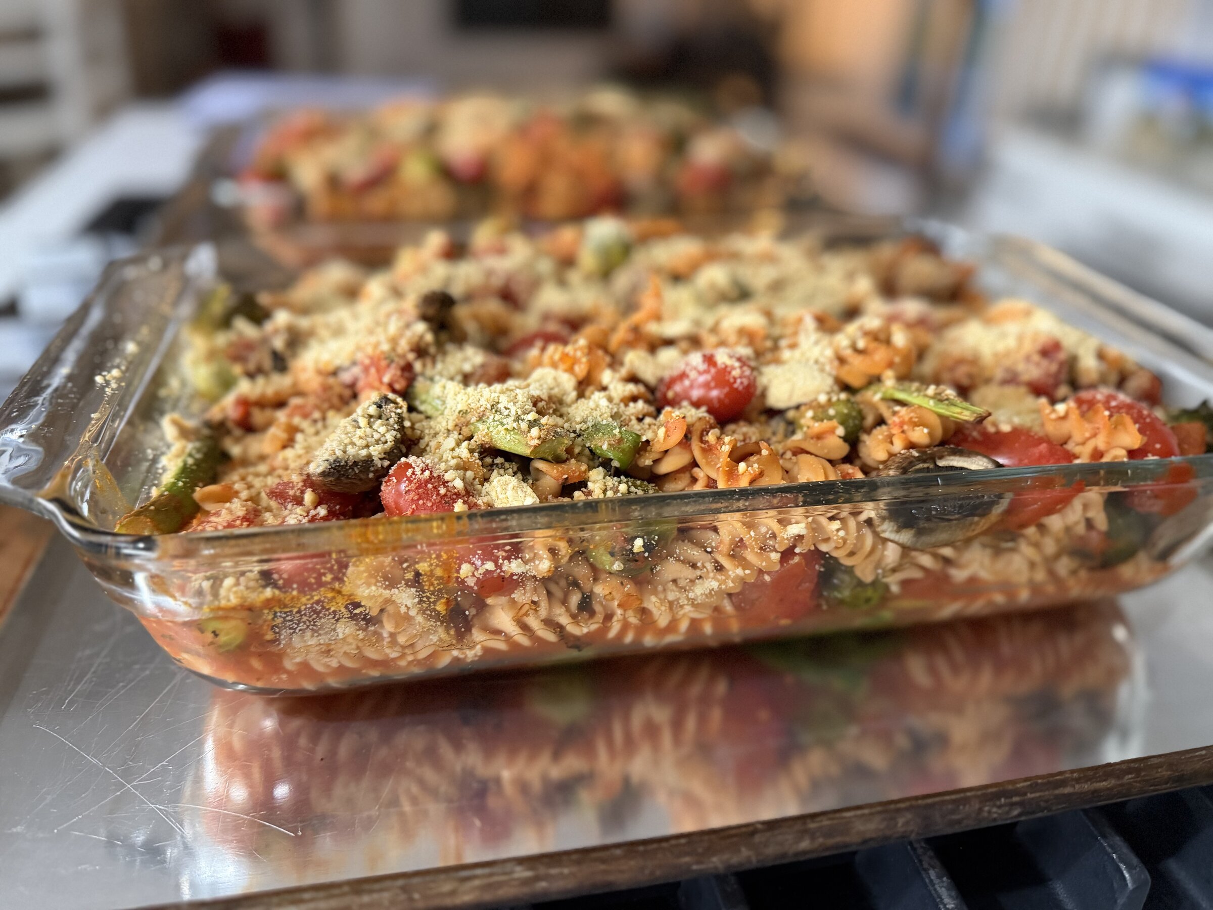Close-up of a glass baking dish of rotini pasta bake topped with roasted asparagus, broccolini, cherry tomatoes, mushrooms, and a dusting of golden cashew parmesan, with a second pan blurred behind it