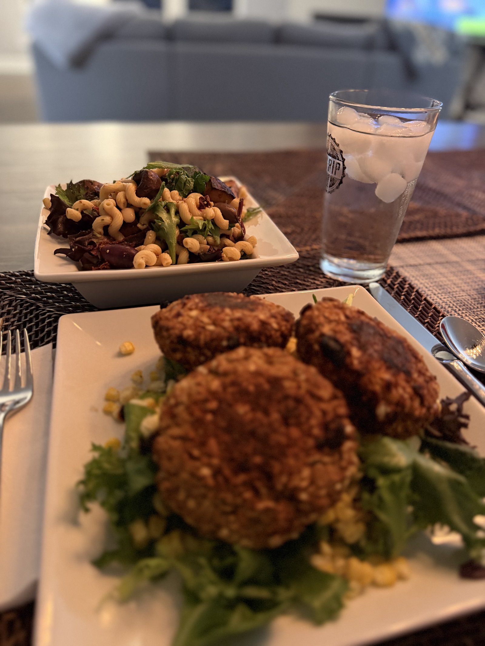 A dinner table set with a plate of cavatappi pasta salad tossed with sun-dried tomatoes, kalamata olives, white beans, and mixed greens, alongside a plate of three seared black bean patties over greens with sweet corn, and a glass of ice water